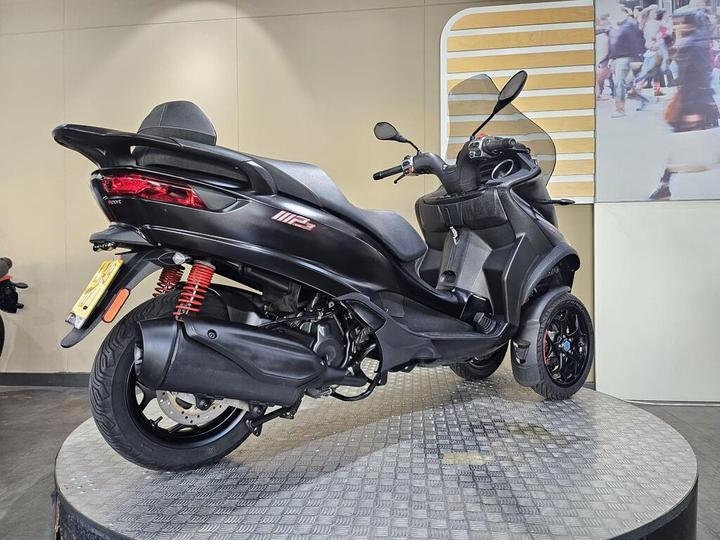 A sleek black three-wheeled scooter with a modern design, featuring red suspension springs and stylish alloy wheels, displayed on a circular platform against a neutral backdrop.