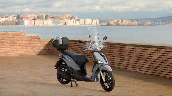 A sleek gray scooter parked on a stone patio, with a blurred waterfront city skyline in the background under a partly cloudy sky.