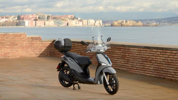 A sleek gray scooter parked on a stone patio, with a blurred waterfront city skyline in the background under a partly cloudy sky.