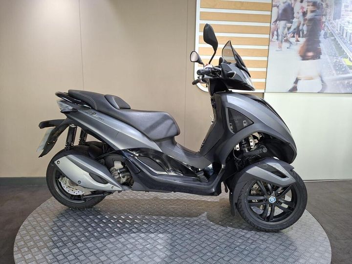 A sleek grey three-wheeled scooter displayed on a circular platform with a simple background.