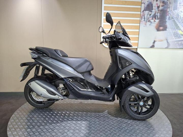 A sleek grey three-wheeled scooter displayed on a circular platform with a simple background.