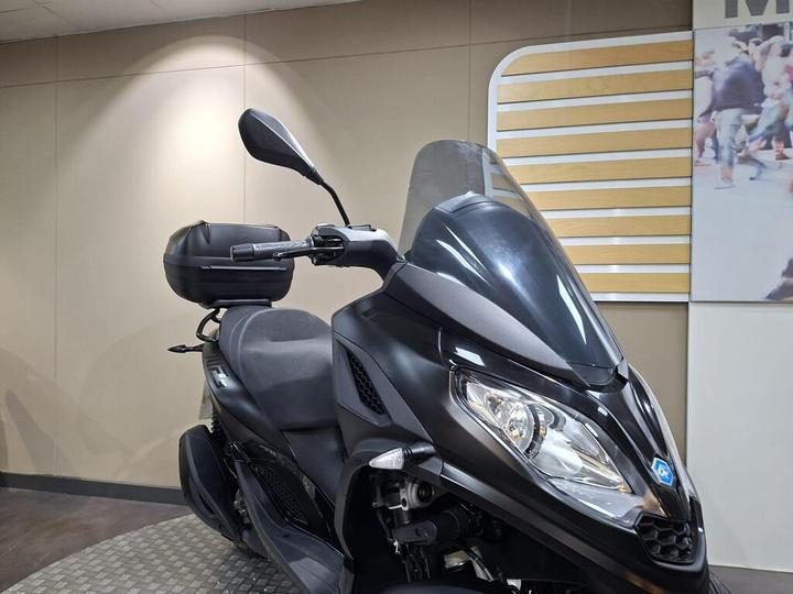 A sleek black scooter with a large windscreen and a rear storage box, positioned on a textured circular tile surface against a minimalist wall background.