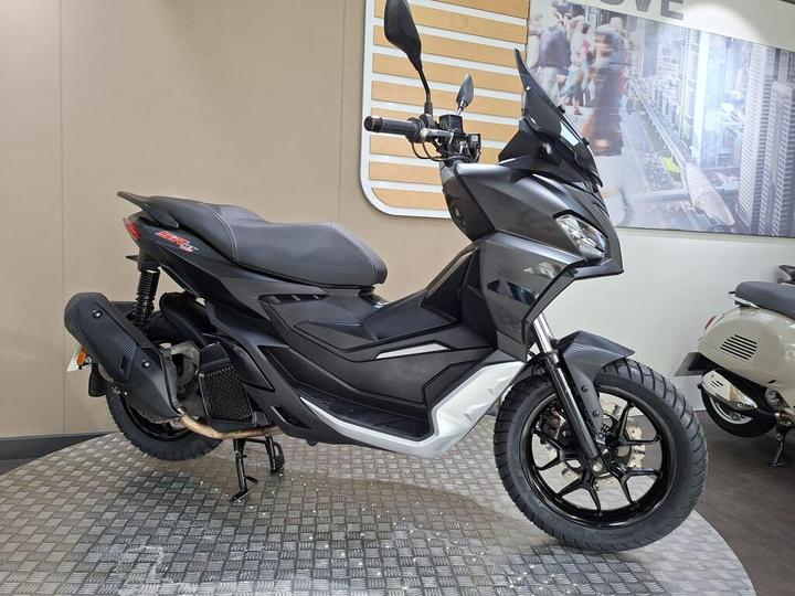 A sleek, modern black scooter displayed in a showroom, featuring a sporty design, front windshield, and textured seating. The background includes a cityscape image and other scooters.
