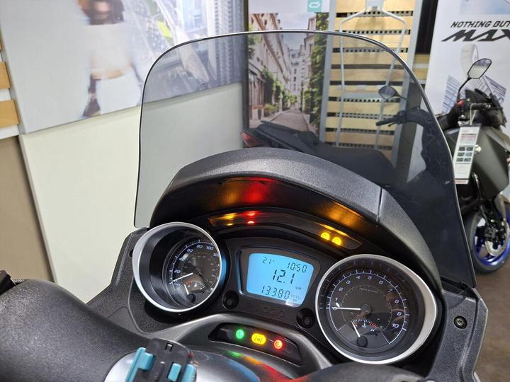 Close-up view of a motorcycle dashboard displaying speedometer and odometer readings, along with warning lights and a digital screen showing temperature and time. A protective windscreen is visible in the foreground, and a blurred background features urban scenery and another motorcycle.