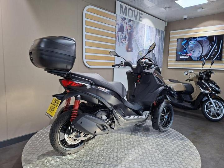 A black three-wheeled scooter with a rear top box is displayed on a circular platform in a showroom, featuring modern decor and a screen showing technical details in the background.