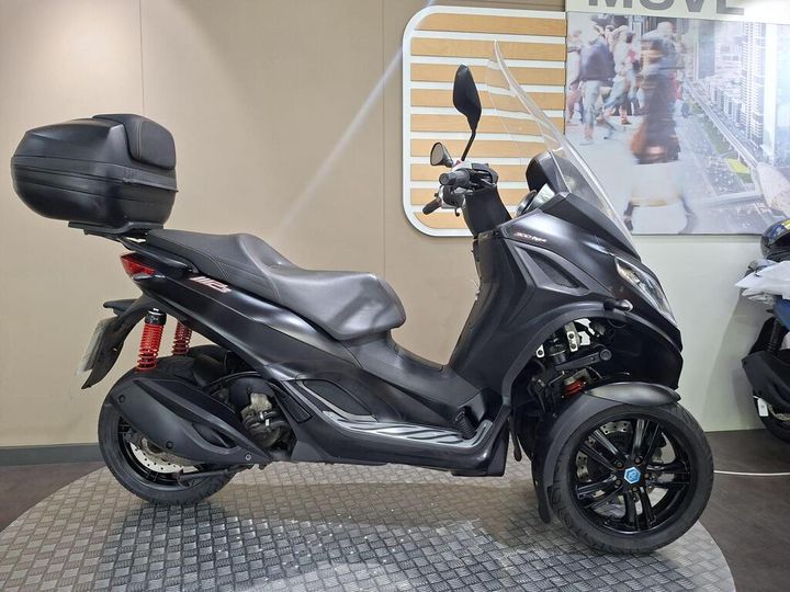 Piaggio MP3 300 HPE Sport 2019 A black three-wheeled scooter with a top case and sleek design, displayed against a light background featuring urban imagery.