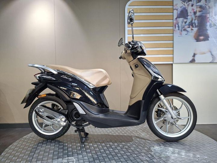 Piaggio Liberty 50 (2020) A sleek black and beige scooter displayed in a showroom, showcasing its modern design and features, with a light-colored wall and an abstract background.