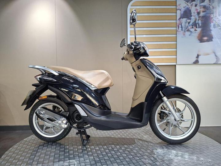 Piaggio Liberty 50 (2020) A sleek black and beige scooter displayed in a showroom, showcasing its modern design and features, with a light-colored wall and an abstract background.