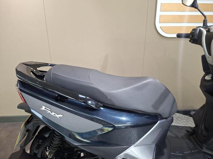 A close-up view of a motorcycle seat and rear section, featuring a textured black seat with red stitching, mounted on a blue motorcycle, set against a beige wall background.