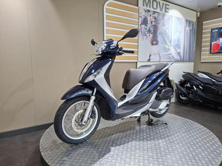 A blue and silver scooter displayed on a circular platform in a showroom, with a modern background featuring people and additional motorcycles.