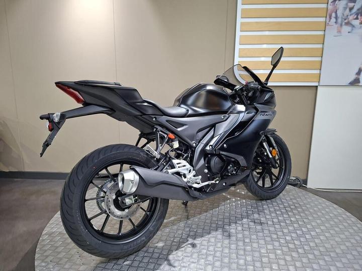 A sleek black motorcycle is positioned on a textured platform, showcasing its rear and side profile. The bike features aerodynamic design elements, a sporty tail, and a round exhaust, set against a neutral-colored wall with wooden accents.