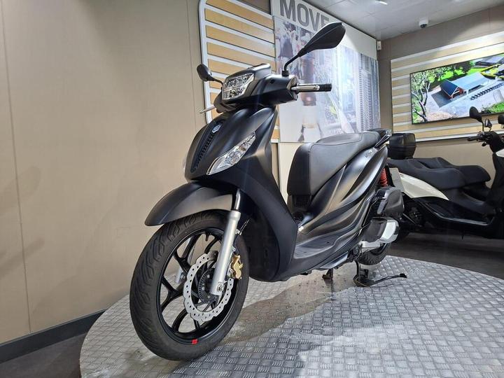 A black scooter displayed on a circular platform in a showroom, with a modern backdrop featuring digital screens and a light-colored wall.