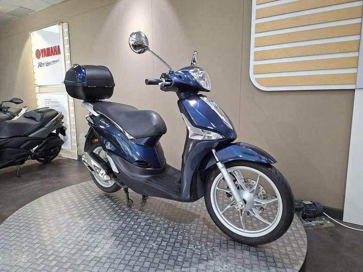 A blue scooter is displayed on a circular platform in a showroom, featuring a comfortable seat, a rear storage compartment, and chrome accents, with a minimalist background.