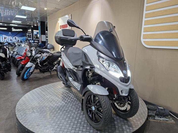 A gray three-wheeled scooter displayed on a circular platform in a motorcycle showroom, with various other bikes visible in the background.