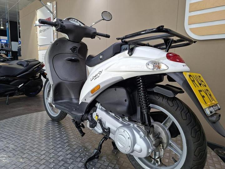 A white scooter parked on a metallic floor, featuring a sleek design with black seating, a rear luggage rack, and a visible license plate. In the background, another scooter is partially visible against a neutral-toned wall.