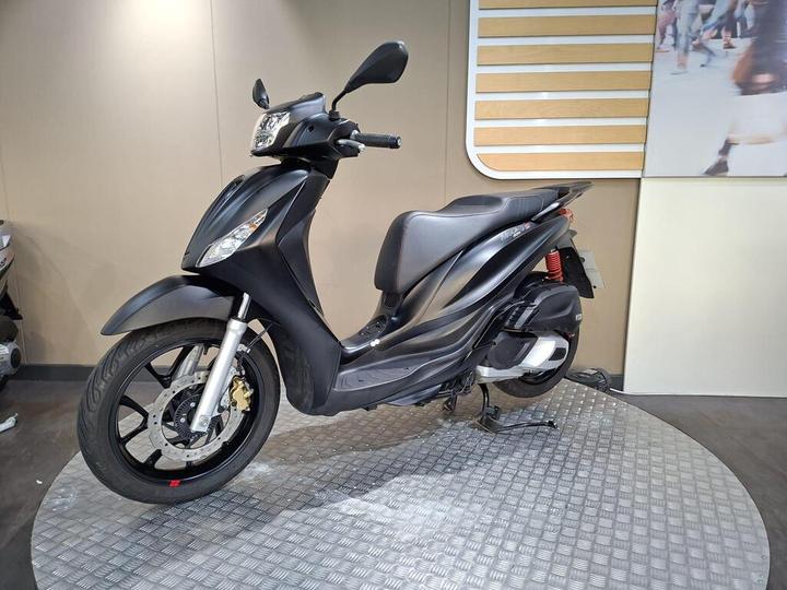 A black scooter displayed on a circular metallic platform against a simple background. The scooter features a sleek design with a prominent front headlight and modern styling.