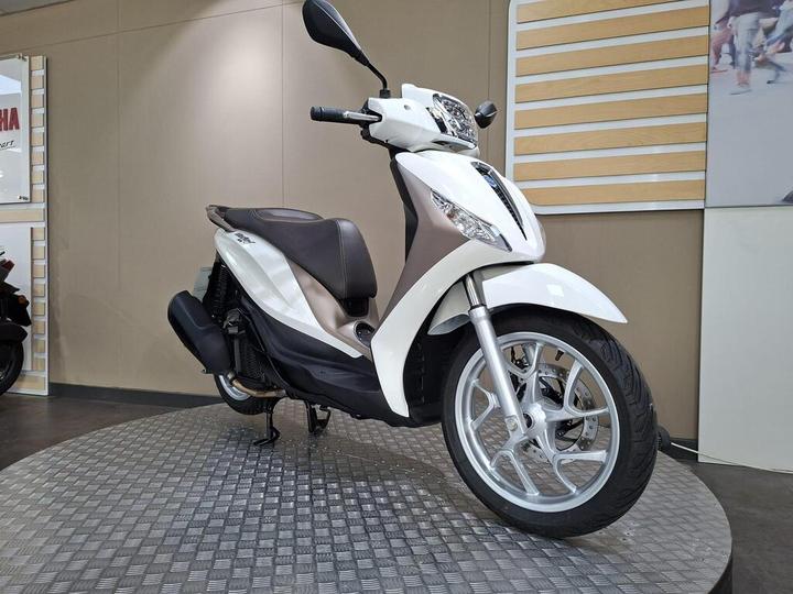 A white and silver scooter displayed on a circular metal platform in a showroom, featuring modern design elements and a detailed front view.