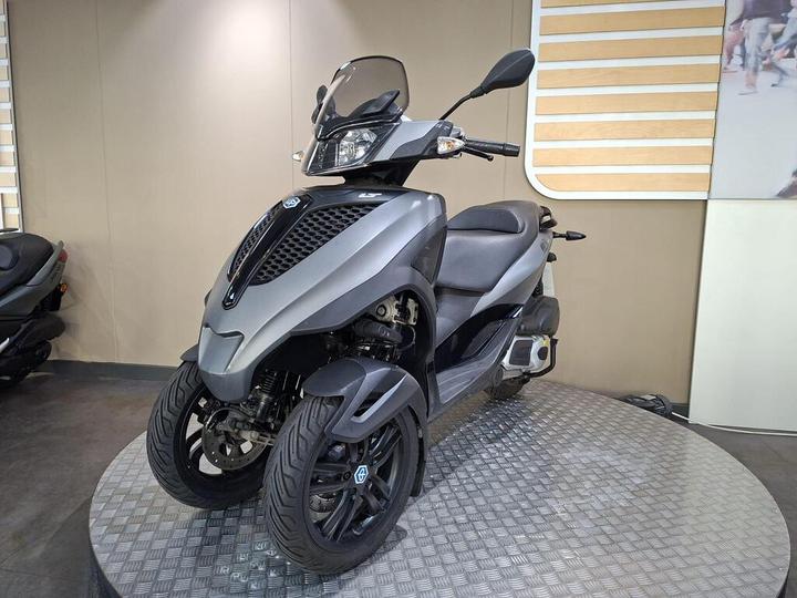 A sleek, modern three-wheeled scooter in metallic gray, parked on a circular platform with a textured surface. The scooter features a windscreen, stylish body design, and two front wheels with prominent tires. The background is neutral, emphasizing the scooter's design.