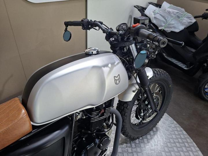 Close-up view of a custom motorcycle featuring a silver fuel tank, brown leather seat, and black handlebars with mirrors, set against a neutral background.