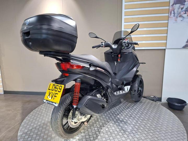 A black scooter with a large rear storage box, displayed on a circular textured platform against a neutral interior backdrop. The scooter features red shock absorbers and is positioned to showcase its rear and side profile.