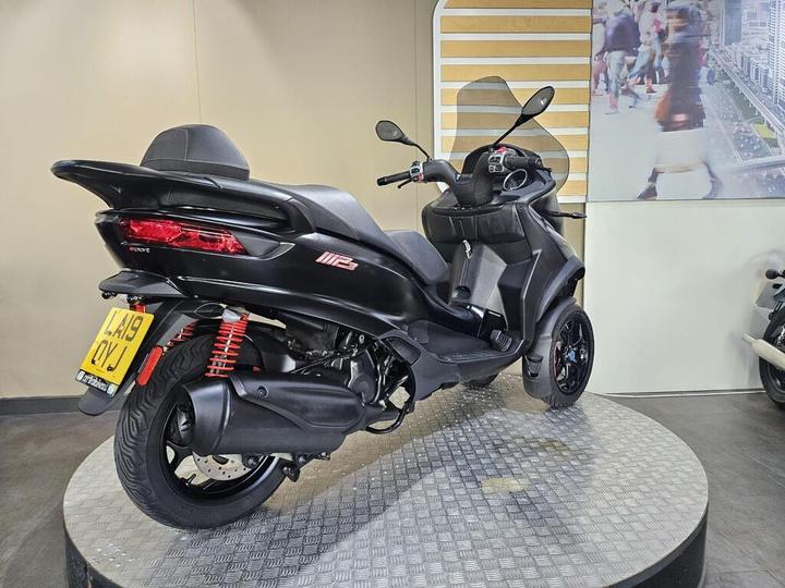 A black three-wheeled scooter displayed on a circular platform, featuring a sleek design, rear red suspension springs, and a license plate visible at the back. The background includes a light-colored wall and a blurred image of a busy street scene.