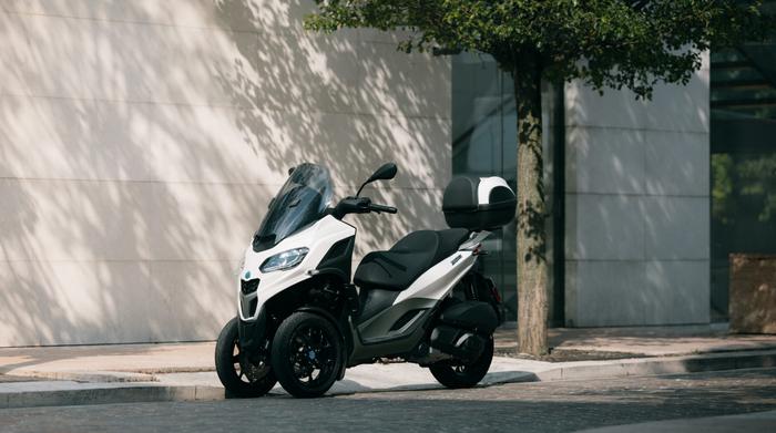 A white three-wheeled scooter parked on a cobblestone street near a modern building, with a tree providing dappled shadow.