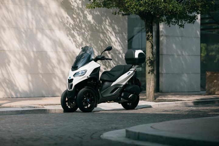 A white three-wheeled scooter parked on a cobblestone street near a modern building, with a tree providing dappled shadow.