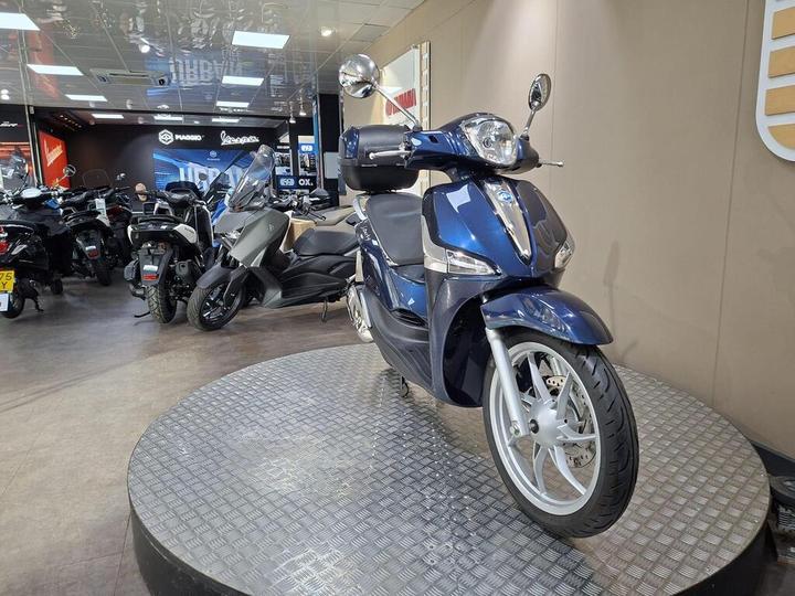 A blue scooter displayed on a round platform in a motorcycle showroom, with various other motorcycles visible in the background.