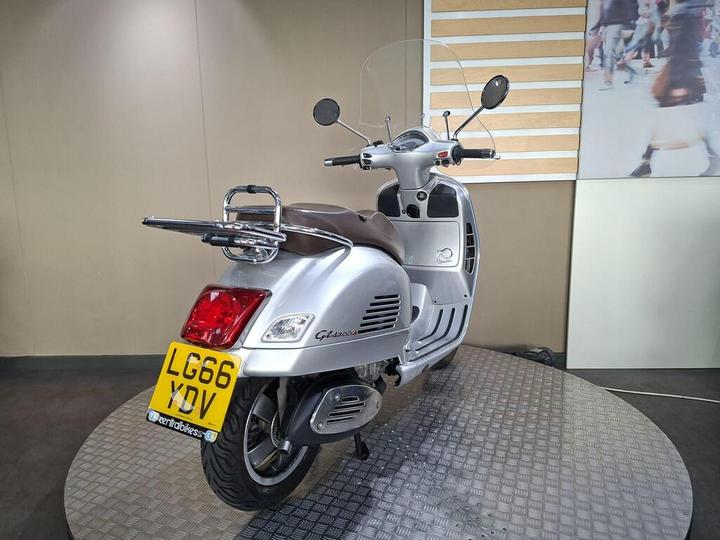 A silver scooter displaying a rear view on a textured circular platform, featuring a brown seat, a windshield, and a license plate reading "LG66 XDV." The background includes a neutral-colored wall and abstract imagery.
