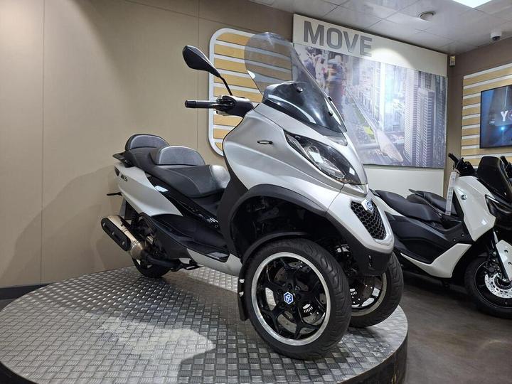 A modern three-wheeled scooter with a sleek silver design is displayed on a circular platform in a showroom. The scooter features a windshield, comfortable seating, and prominent black wheels. In the background, there are images of a cityscape and another scooter.