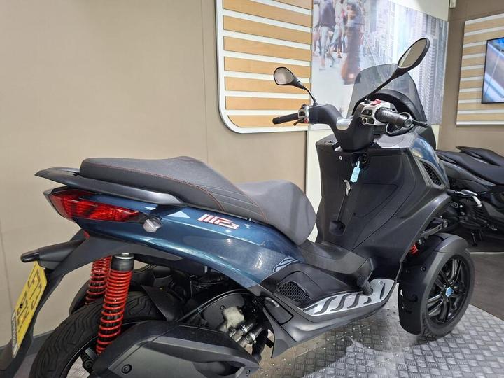 A blue and gray three-wheeled scooter displayed in a showroom, featuring a sleek design, red shock absorbers, and a modern dashboard. The background includes a wall with vertical wooden slats and an image of a cityscape.