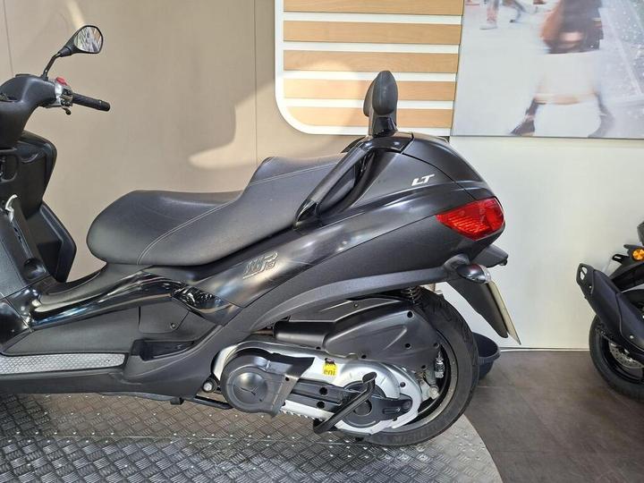 A side view of a black scooter with a sleek design, featuring a visible seat, a rear light, and a silver exhaust. The background displays a light-colored wall with horizontal wooden slats.