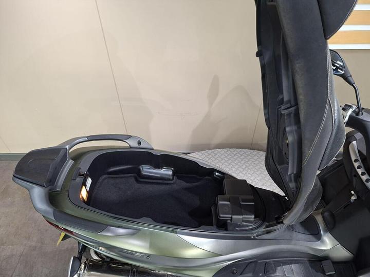 A close-up view of an open motorcycle seat, revealing the storage compartment beneath, featuring a black interior and a small light on the side. The seat is padded and gray, with the motorcycle's body in a metallic green color in the foreground.