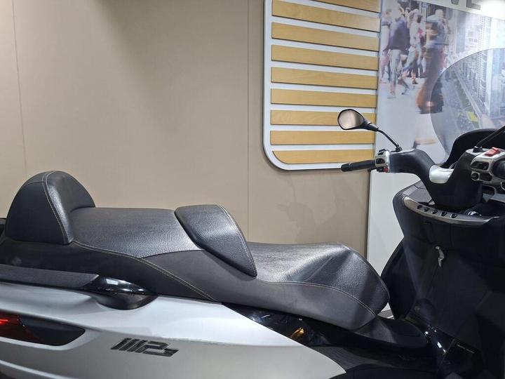 A close-up view of a modern scooter's seat and handlebars, set against a neutral wall with a patterned background.