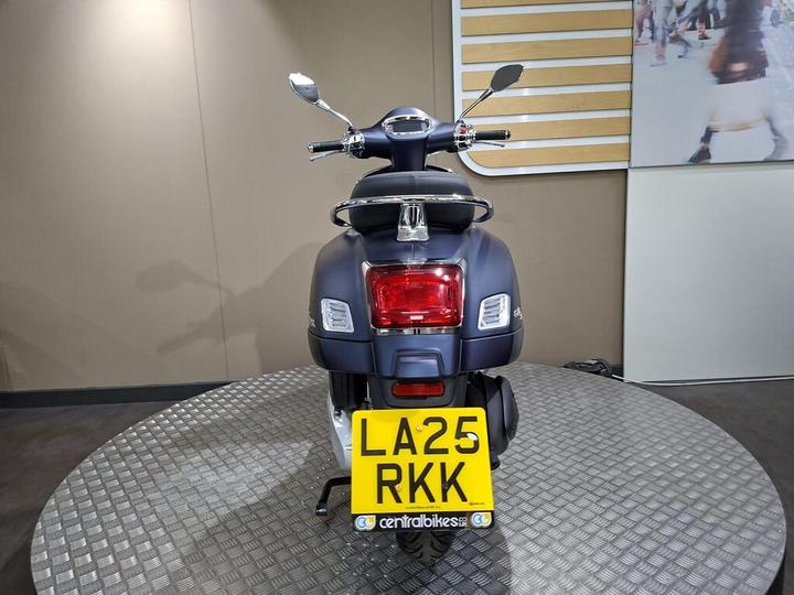 A rear view of a blue scooter on a circular platform, showcasing its sleek design and yellow license plate with the registration "LA25 RKK." The background features a neutral-colored wall and a display of people in motion.