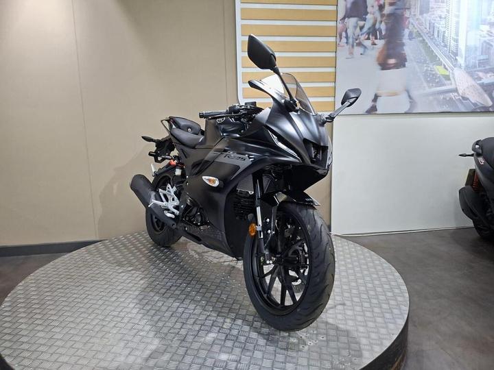 A sleek black motorcycle is displayed on a circular platform, showcasing its aerodynamic design and glossy finish. The background features a minimalistic wall with angled lines and an image of a bustling urban scene.