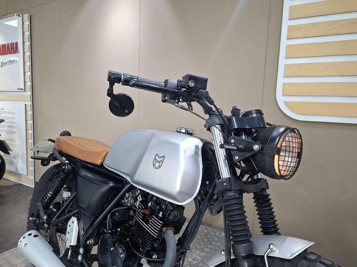A close-up view of a custom motorcycle featuring a silver fuel tank, brown leather seat, and a large round headlight with a grid pattern. The handlebars include a mirror and technical components, with a minimalist design and rugged tires visible in the foreground. The backdrop includes Yamaha branding and promotional materials.