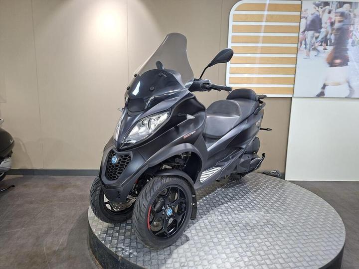 A black three-wheeled scooter featuring a prominent windshield, displayed on a circular platform with a textured surface, against a neutral backdrop.