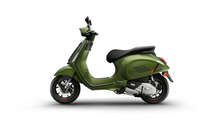 A green Vespa scooter parked on a white background, shown in profile with its distinctive round body, flat seat, and vintage-style features.