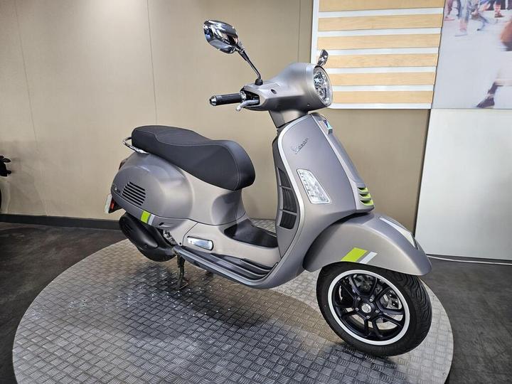 A silver-grey scooter with a black seat and sporty black wheels displayed on a circular platform with a textured surface against a beige wall.