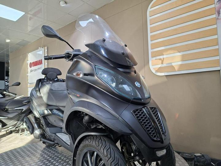 A black three-wheeled scooter with a windshield parked indoors, partially visible behind another motorcycle, with a Yamaha logo and branded signage in the background.