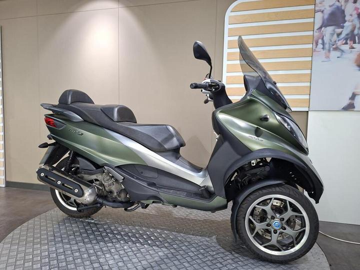 A green and silver three-wheeled scooter displayed on a textured circular platform against a neutral background.