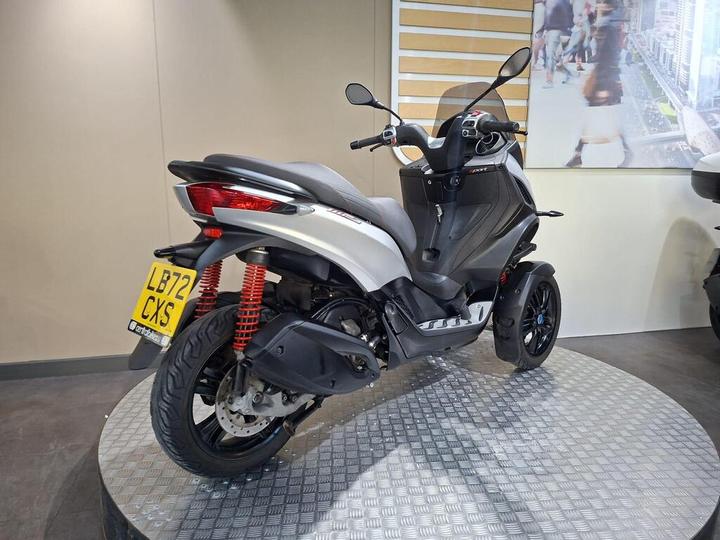 A gray and black three-wheeled scooter is displayed on a circular metallic platform, showcasing its sleek design and features, including red shock absorbers and a license plate reading "LB72 CXS." The background features a light-colored wall and an image of people walking in an urban setting.