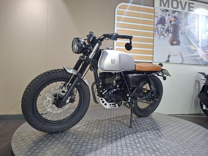 A vintage-style motorcycle with a silver tank and brown leather seat, featuring rugged tires and a distinctive round headlight, displayed on a textured platform against a neutral background.