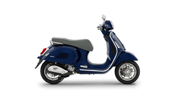 A blue Vespa scooter with a grey seat, viewed from the side against a white background.