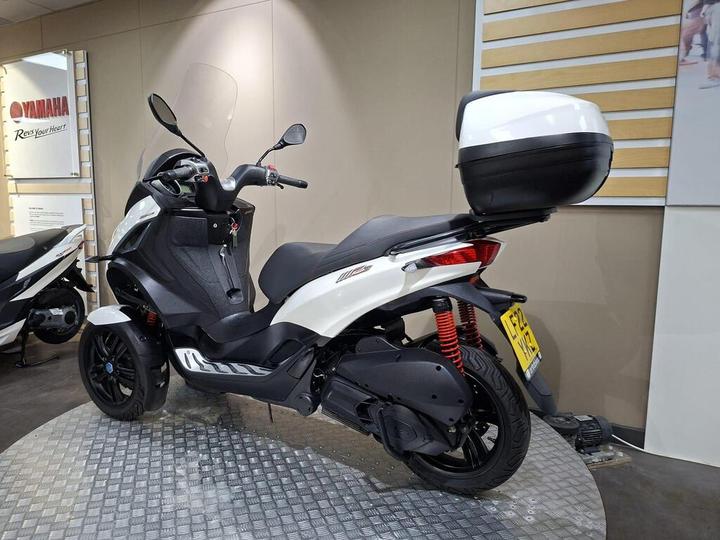 A white and black three-wheeled scooter parked on a textured surface, displaying a storage compartment at the rear, with a Yamaha logo visible in the background.