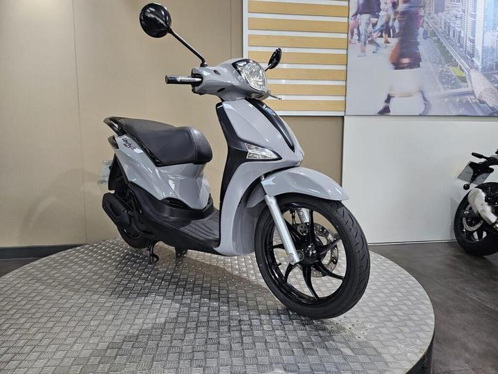 A sleek grey scooter displayed on a circular metal platform, with a blurred background featuring urban scenery and other scooters.