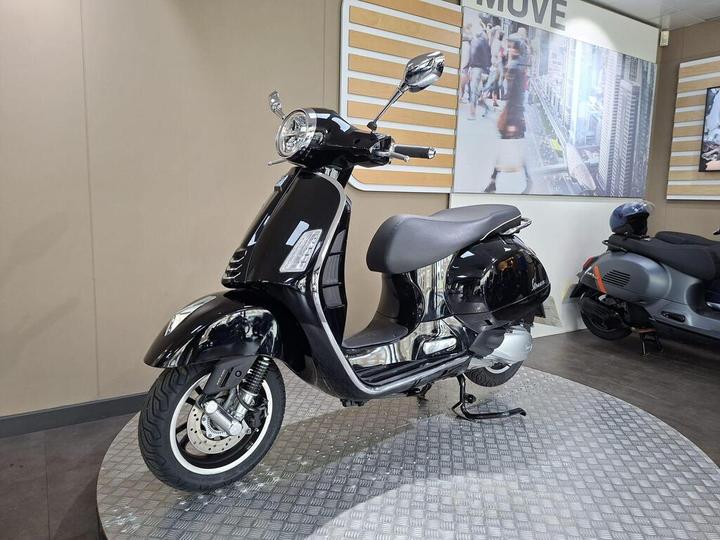A sleek black scooter displayed on a circular stand in a showroom, with modern decor featuring an urban-themed background and another scooter partially visible in the background.