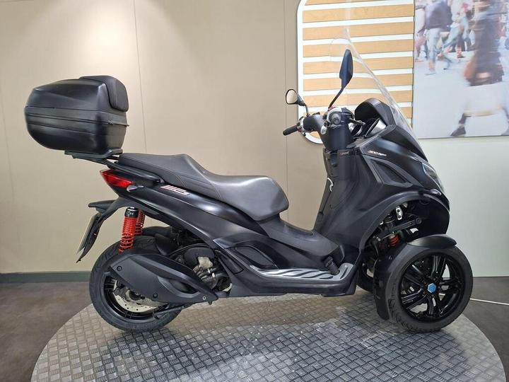 A black three-wheeled scooter with a top case, featuring a sleek design and prominent rear suspension, displayed on a textured circular platform against a neutral background.