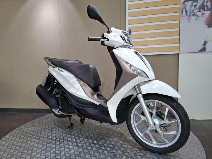A white and silver scooter displayed on a circular metallic stand with a textured surface, set against a neutral-colored backdrop with vertical wooden panels.